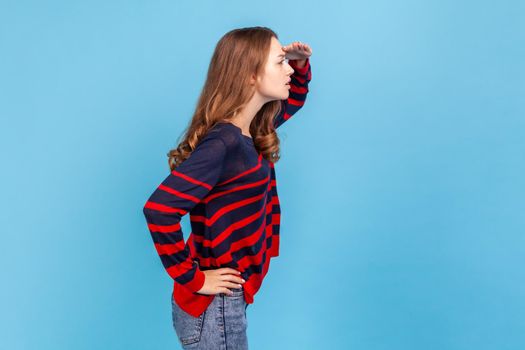 Side view portrait of woman wearing striped casual style sweater keeps hand near forehead looks far away searches something on horizon. Indoor studio shot isolated on blue background.