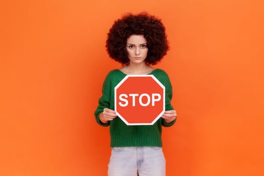 Serious strict woman with Afro hairstyle wearing green casual style sweater holding red stop sign in hands, looking at camera, forbidden actions. Indoor studio shot isolated on orange background.