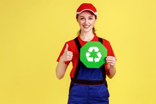 Portrait of optimistic worker woman standing and holding recycling sign, thinking green, showing thumb up, wearing overalls and red cap. Indoor studio shot isolated on yellow background.