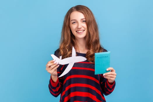 Smiling woman wearing striped casual style sweater, showing paper plane and passport and dreaming of vacation abroad, trip around world. Indoor studio shot isolated on blue background.