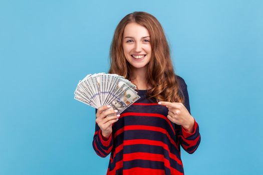 Optimistic woman wearing striped casual style sweater, pointing finger and big fan of dollars banknotes, looking at camera with satisfied expression. Indoor studio shot isolated on blue background.