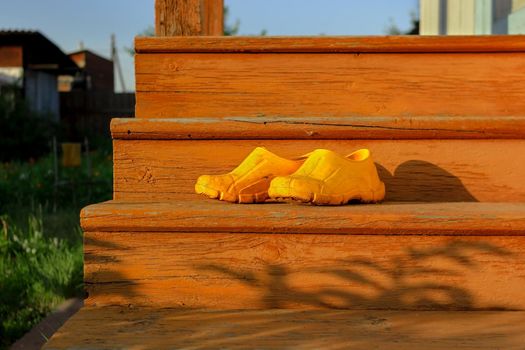 Yellow rubber muddy work shoes on old orange stairs on summer backyard at sunset, village and countryside authentic mood, low angle