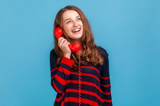 Young adult woman wearing striped casual style sweater, talking via red retro landline phone, having pleasant conversation, looking away with smile. Indoor studio shot isolated on blue background.