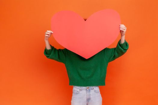 Unknown woman wearing green casual style sweater holding big red paper heart, hiding behind love huge love symbol, shy to express her feelings. Indoor studio shot isolated on orange background.