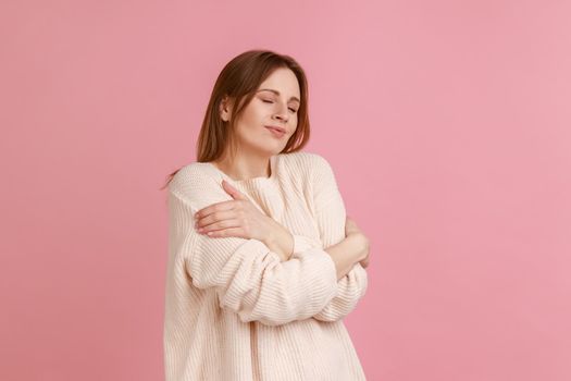 I love myself. Portrait of woman hugging herself and smiling, feeling comfortable and fulfilled, narcissistic egoistic person, wearing white sweater. Indoor studio shot isolated on pink background.