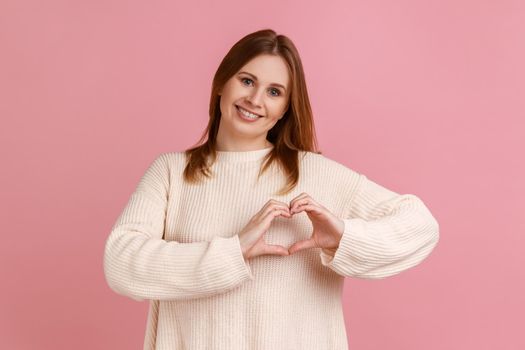 Portrait of blond woman making heart shape with fingers, gesturing love hope charity sign, looks at camera, wearing white sweater. Indoor studio shot isolated on pink background.