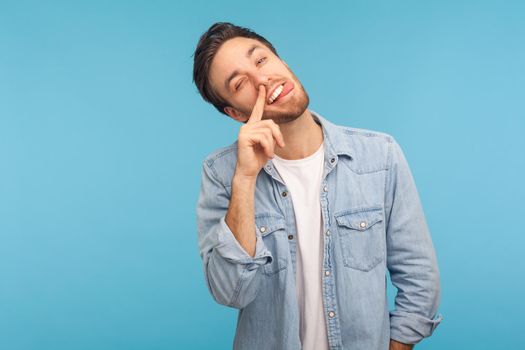 Portrait of funny man wearing denim shirt, putting finger into his nose and showing tongue, fooling around, bad habits, disrespectful behavior. Indoor studio shot isolated on blue background.