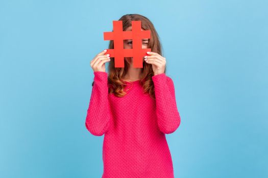 Portrait of attractive young adult woman wearing pink pullover, looking through big red hashtag symbol, popular internet idea, viral content. Indoor studio shot isolated on blue background.