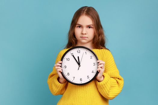 Portrait of little girl holding big wall clock in hands, looking at camera with serious expression, wearing yellow casual style sweater. Indoor studio shot isolated on blue background.