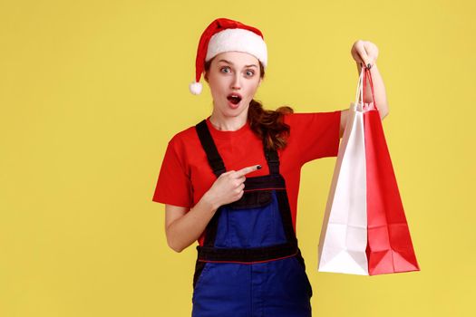 Shocked delivery woman holding pointing at shopping bags, offering free shipping from fashion store, wearing blue overalls and santa claus hat. Indoor studio shot isolated on yellow background.