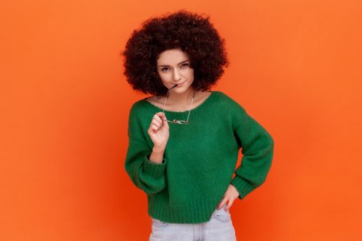 Pensive woman with Afro hairstyle wearing green casual style sweater standing holding glasses in and, thinking about new plan, looking at camera. Indoor studio shot isolated on orange background.