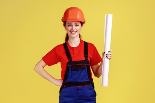 Portrait of smiling delighted woman builder holding blueprints, keeps hand on hip, looking at camera, wearing overalls and protective helmet. Indoor studio shot isolated on yellow background.