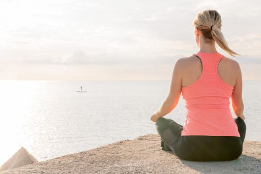 back of an unrecognizable blonde woman practicing yoga at sunrise sitting in the lotus position in front of the sea, concept of mental health and relaxation, copy space for text