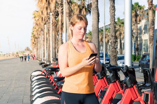 attractive blonde girl looking at the phone next to a rental bike park on the city's promenade, concept of sport and healthy lifestyle