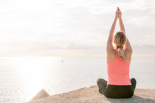 back view of an unrecognizable blonde woman practicing yoga at sunrise sitting in front of the sea, concept of mental health and relaxation, copy space for text