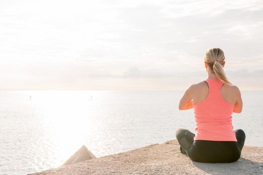 back view of an unrecognizable blonde woman doing yoga at dawn sitting in front of the sea, mental health care and relaxation concept, copy space for text