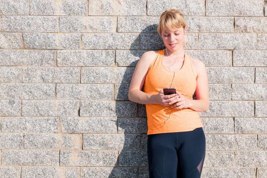 young sporty caucasian girl with earphones looking at her mobile phone leaning against a stone wall, technology concept and healthy lifestyle, copy space for text