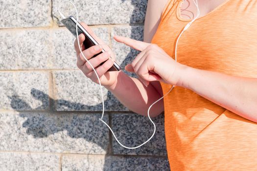 hands of a young sporty girl with earphones touching her mobile phone's screen leaning against a stone wall, technology and healthy lifestyle concept