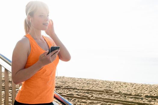young sporty blonde woman with earphones listening to music from her mobile phone by the beach, technology and healthy lifestyle concept, copy space for text and lens flare effect
