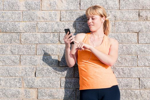young sporty blonde girl with earphones consulting her mobile phone leaning against a stone wall, technology concept and healthy lifestyle, copy space for text