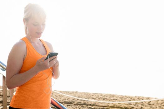 young sporty blonde woman with earphones using her mobile phone by the beach, technology concept and healthy lifestyle, copy space for text and lens flare effect