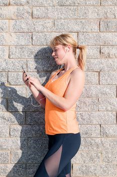 vertical photo of a young sporty blonde girl with earphones consulting her mobile phone leaning against a stone wall, technology concept and healthy lifestyle, copy space for text