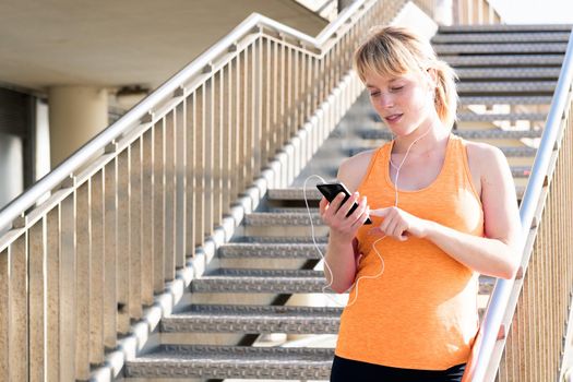 young blonde woman with earphones using her mobile phone next to a staircase, concept of technology and healthy lifestyle