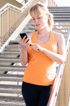 vertical photo of a young blonde woman with earphones using her mobile phone next to a staircase, concept of technology and healthy lifestyle