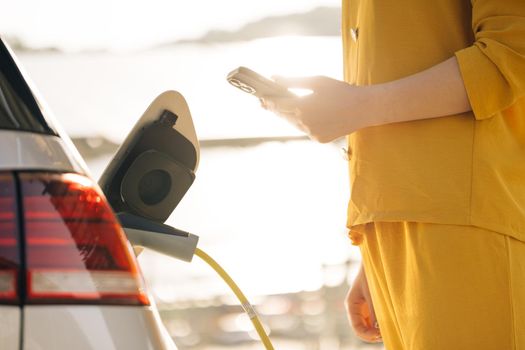 Business woman connects plug to electric car activates charging app on phone. Girl plugs power cable to charge electric car in parking lot. Eco friendly alternative energy green environment concept.