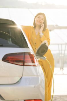 Positive woman have talking conversation by phone near her electric car and waits when the vehicle will be charged. Confident business woman talking on phone near car outdoors.