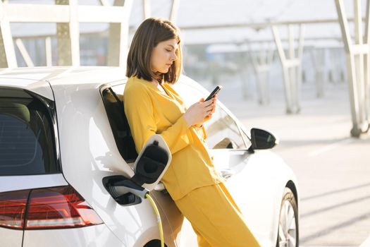 Caucasian woman stands with mobile phone near her electric car and waits when vehicle will charged. Charger plug of an electric car. Electric vehicle charging near an solar power plant