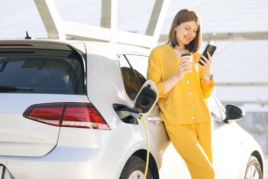 Caucasian woman using smart phone and waiting power supply connect to electric vehicles for charging the battery in car. Plug charging an Electric car from charging station.