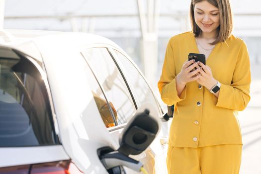 Caucasian woman use smart phone while waiting and power supply connect to electric vehicles for charging the battery in car. Electric vehicle charging near an solar power plant