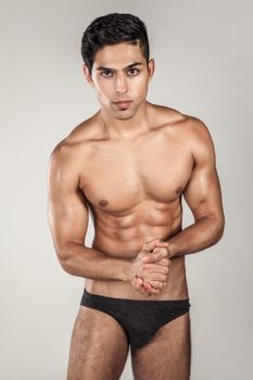 Portrait of handsome sexy brunette man with tanned body, demonstrating his perfect sporty body, looking at camera, wearing black panties. Indoor studio shot isolated on gray background.