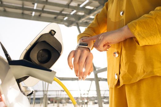 Businesswoman plugging in charging cable to to electric vehicle. Female hand inserts power connector into EV car and charges batteries, uses smartwatch for activates start charging