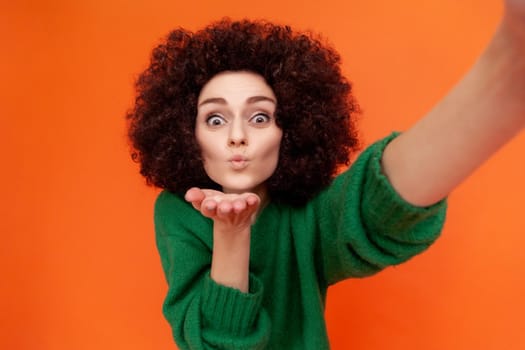 Positve friendly woman with Afro hairstyle in green casual style sweater making selfie POV, sending air kisses to followers while having livestream. Indoor studio shot isolated on orange background.