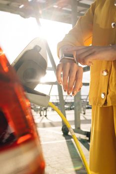 Close up of woman hands attaching power cable supply to charge electric or EV car using app on wearable smart watch. Female plugging an electric car or EV at electric charging station.
