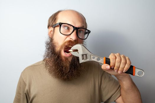 Funny male pulling out tooth with wrench, having foolish expression, looks away with crossed eyes, bald bearded man wearing T-shirt and glasses. Indoor studio shot isolated on gray background.