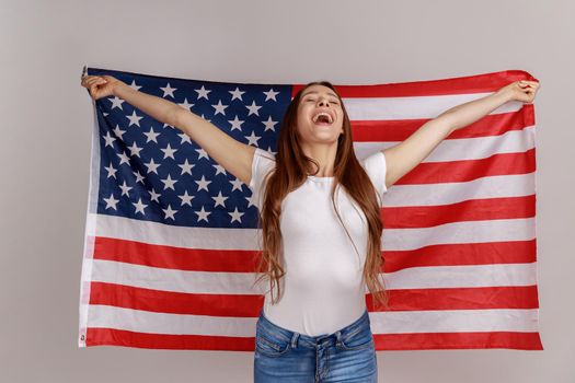 Portrait of attractive beautiful young woman with dark hair, holding USA flag over shoulders, rejoicing happily, wearing white T-shirt. Indoor studio shot isolated on gray background.