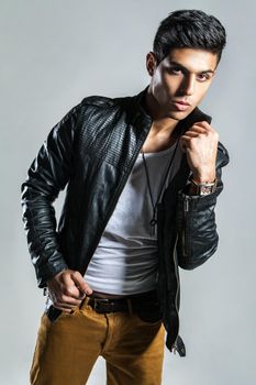 Portrait of sexy confident attractive man wearing fashionable leather jacket, keeping hand on his collar, looking at camera with confident expression. Indoor studio shot isolated on gray background.