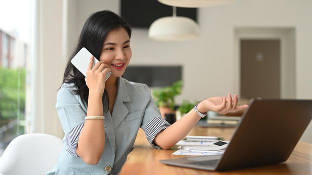 Asian female employee having pleasant phone conversation and using laptop at office desk.