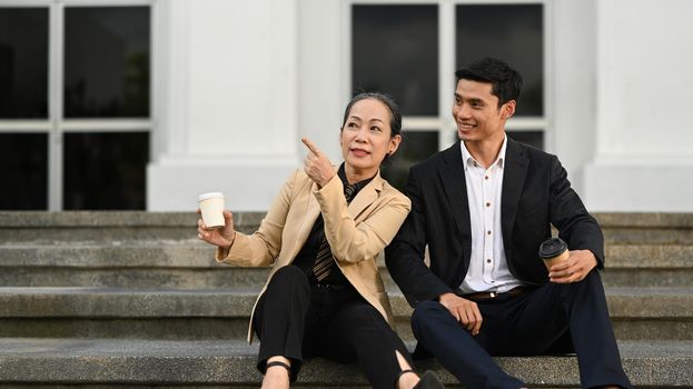 Mature businesswoman and young businessman talking each other, sitting on the stairs in front of business center.
