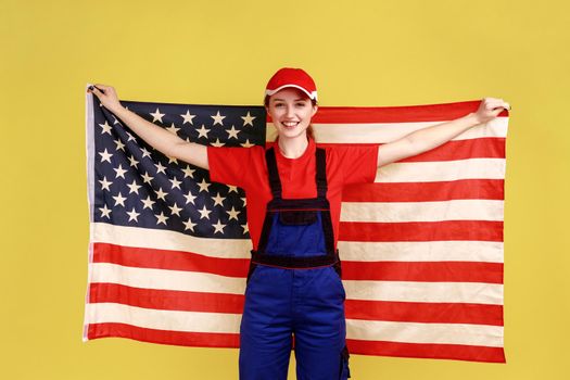 Portrait of happy smiling worker woman standing with american flag, celebrating national holiday, looking at camera, wearing overalls and red cap. Indoor studio shot isolated on yellow background.