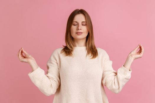 Portrait of blond woman closing eyes and holding hands up in mudra gesture, meditating, yoga exercise to reduce stress, wearing white sweater. Indoor studio shot isolated on pink background.