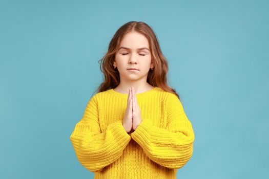 Portrait of little girl holding fingers in mudra gesture, feeling calm positive and relaxed, yoga practice, wearing yellow casual style sweater. Indoor studio shot isolated on blue background.