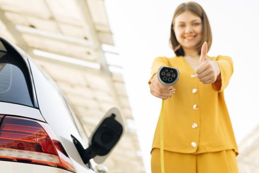 Female showing thumbs up holding power cable supply plugged at electric car charging station. Woman standing near electric charging station looking at camera and showing thumb up.