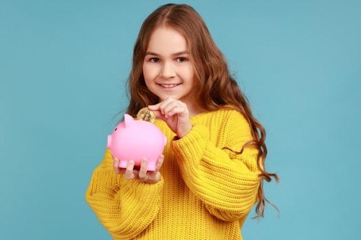 Portrait of little girl putting golden crypto coin into piggy bank, looking at camera with smile, wearing yellow casual style sweater. Indoor studio shot isolated on blue background.