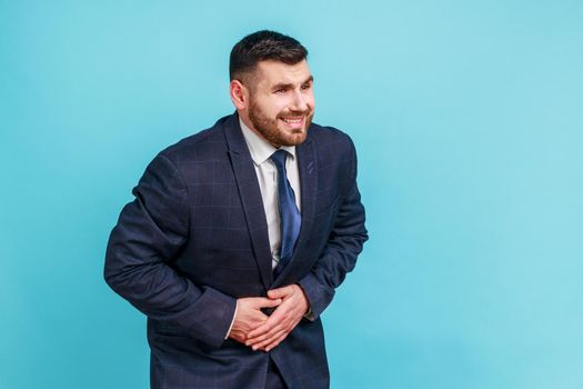 Sick bearded businessman wearing official style suit holding hands on stomach feeling acute pain, suffering indigestion and nausea, duodenal ulcer. Indoor studio shot isolated on blue background.