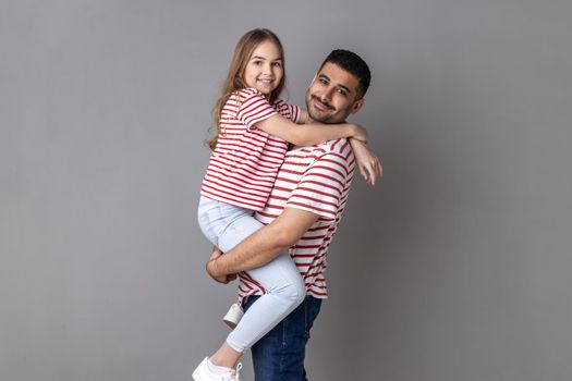 Portrait of happy positive father and daughter in striped T-shirts standing looking at camera with happy facial expressing, daddy holding kid. Indoor studio shot isolated on gray background.