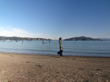 Man wearing a hoodie, pants, and shoes Handstands at on beach in Sausalito is a San Francisco Bay Area city in Marin County, California with boats in bay and Angel Island in background. 